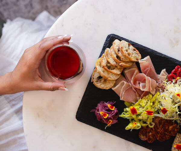 A hand holds a glass of red beverage near a charcuterie board with bread, meat, and colorful garnishes on a table.