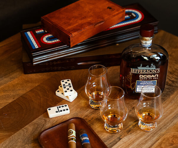 The image shows cigars on a tray, a bottle of whiskey, three glasses, dominoes, and board game pieces on a wooden table.