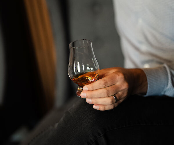 A person holding a glass of whiskey while sitting on a sofa, wearing a light shirt and dark pants, with a wood-panel background.