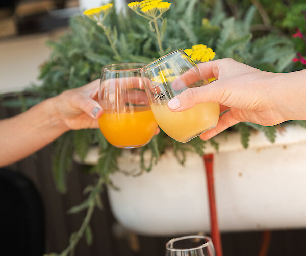 Three hands holding glasses with orange drinks, toasting. A glass with red drink is on the table. Yellow flowers in the background.
