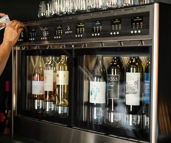 A wine dispensing machine with several bottles and multiple taps, surrounded by glasses and colorful bottle labels on top shelves.