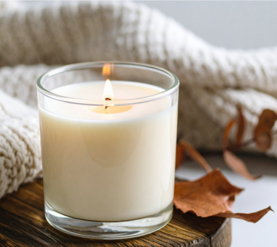 A lit candle in a glass jar sits on a wooden surface with a knitted blanket and dried leaves in the background.