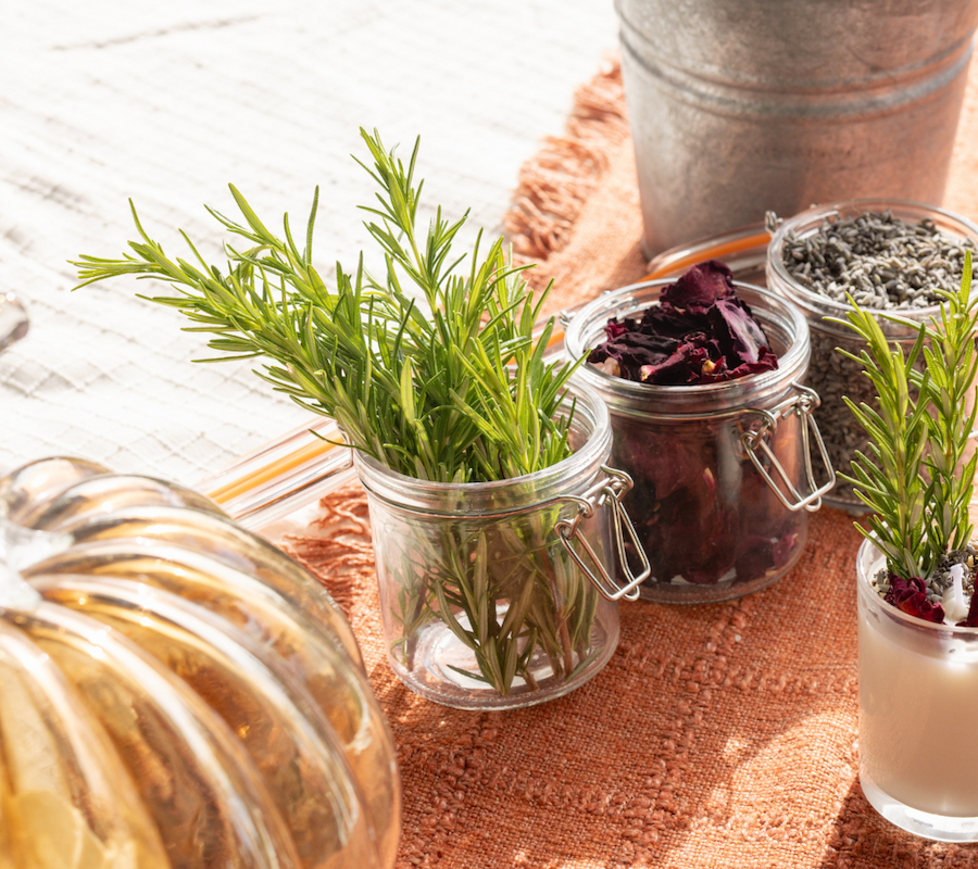 Jars with rosemary, dried flowers, and lavender on a textured cloth, next to a glass pumpkin and a small bottle.