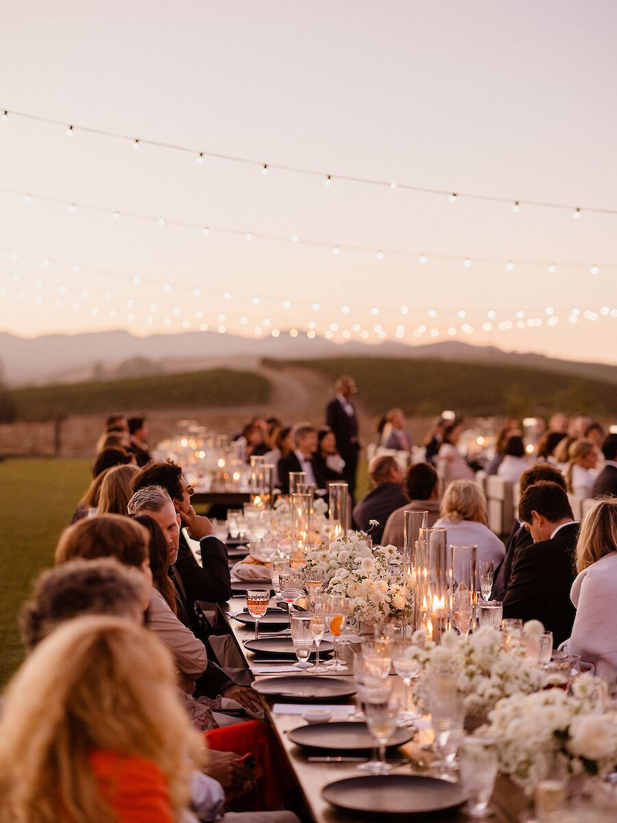 People are seated at long tables outdoors under string lights, surrounded by fields, with flowers and candles decorating the tables.