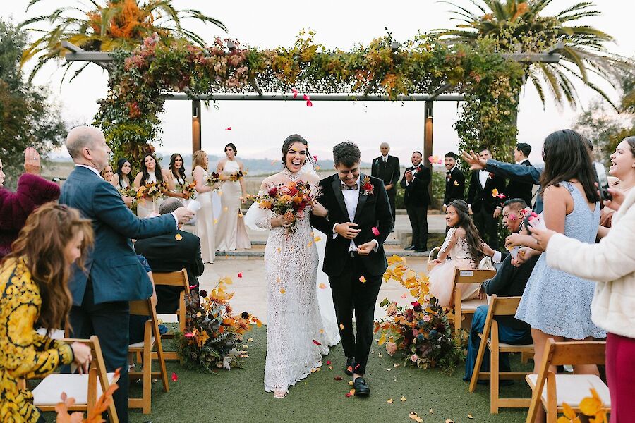 A bride and groom walk down the aisle outdoors, surrounded by cheering guests and floral decorations.