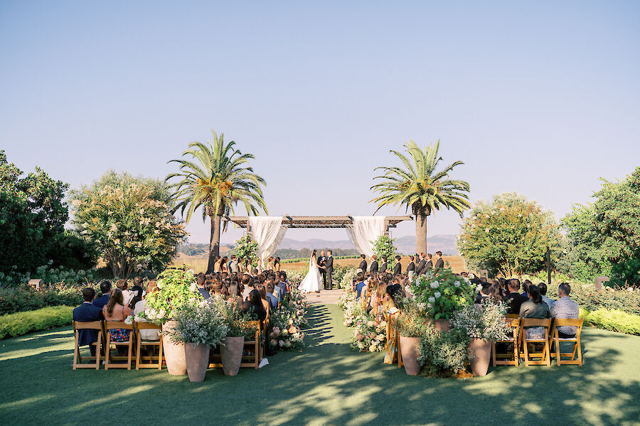 Outdoor wedding ceremony with guests seated on either side of a grassy aisle, surrounded by greenery and palm trees, under clear skies.