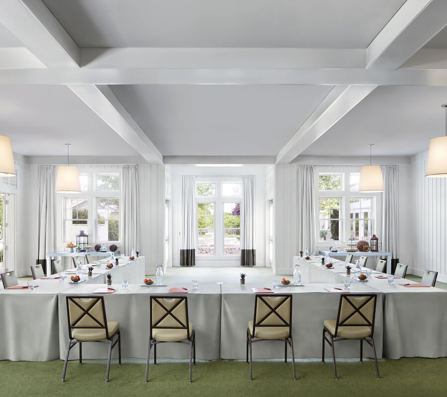 A bright conference room with a U-shaped table, chairs, hanging lights, and large windows letting in natural light.