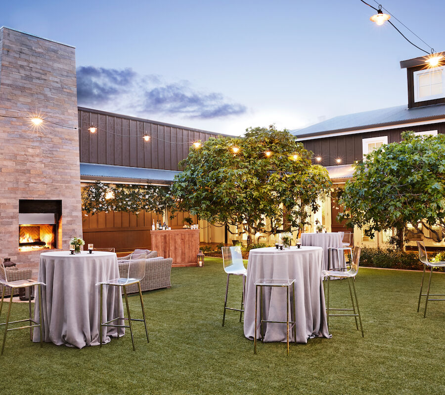 An outdoor space with round tables covered in gray cloth, bar stools, trees, string lights, and a fireplace in the background.