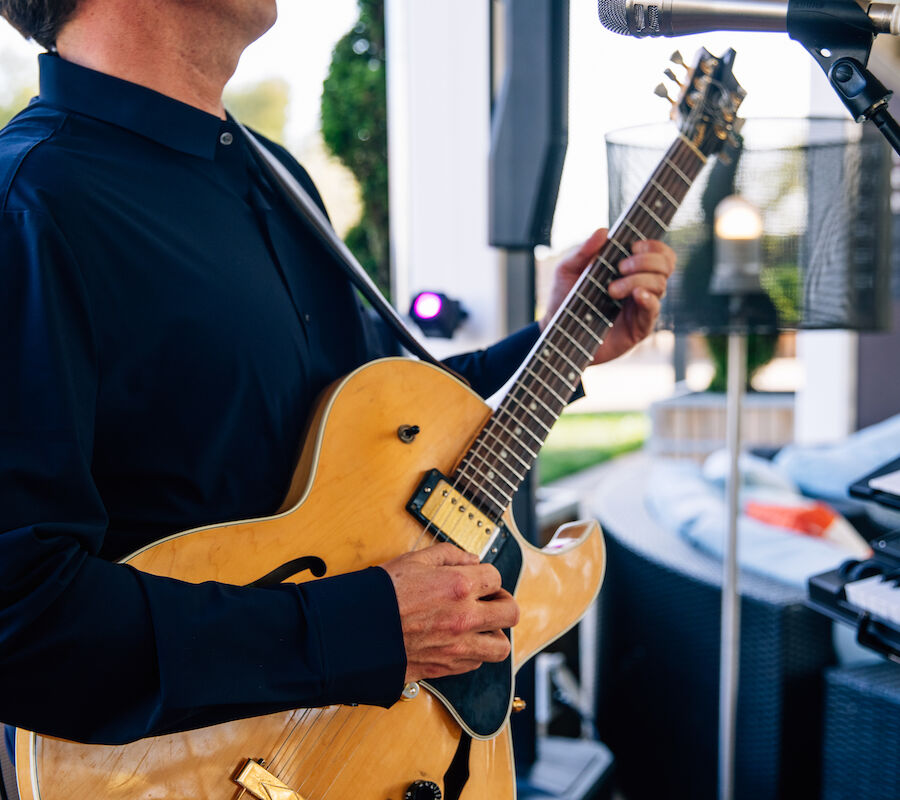 A person in a dark shirt plays a yellow electric guitar, standing near a microphone, with a keyboard visible in the background.