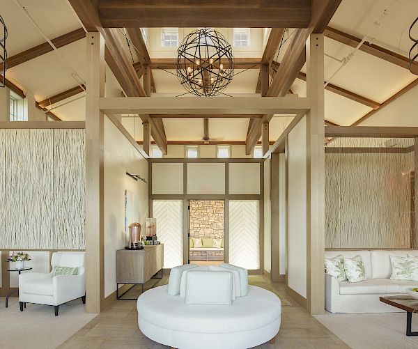 A spacious living area with modern decor, featuring white seating, light wood accents, geometric chandeliers, and high ceilings for a bright, airy feel.