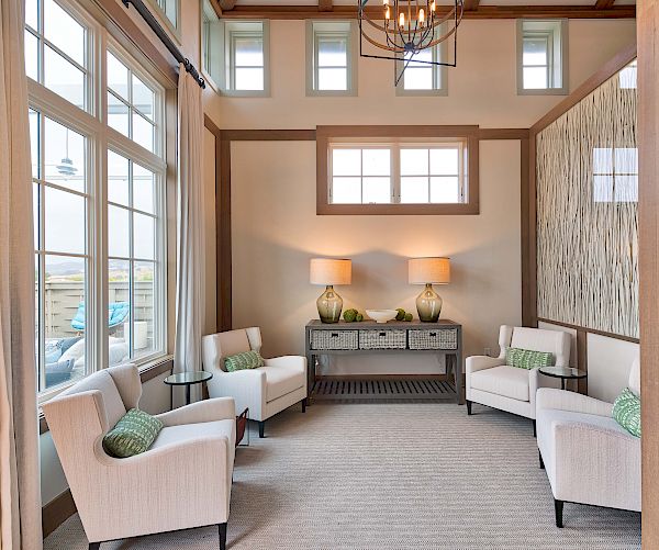 A cozy sitting room featuring four white armchairs with green pillows, a side table, a console with two lamps, and large windows bringing in natural light.