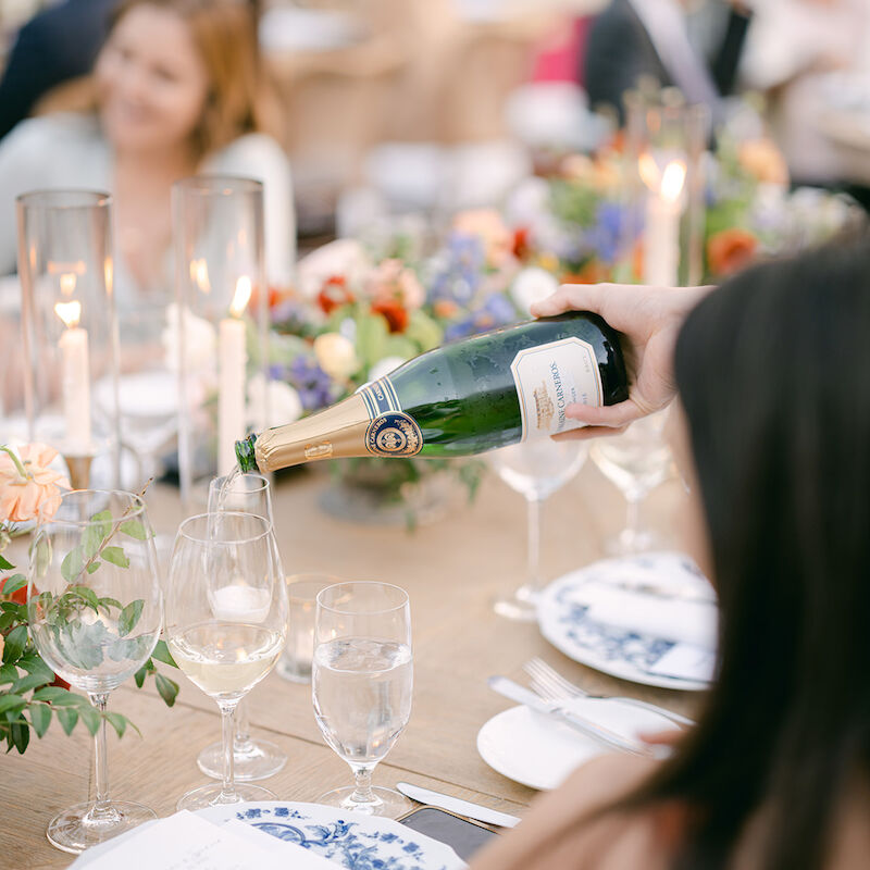 A person is pouring champagne into a glass at a table set for a formal event, with flower arrangements and tableware visible.