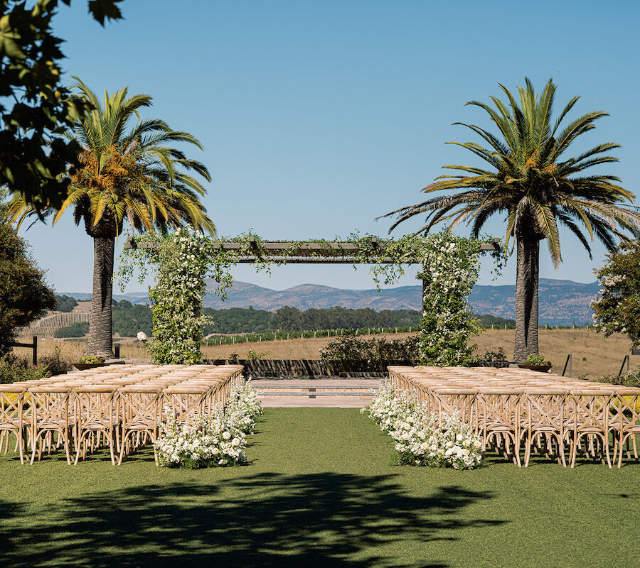 An outdoor wedding setup features rows of wooden chairs facing a green arch adorned with foliage. Palm trees, mountains, and clear skies surround the scene.