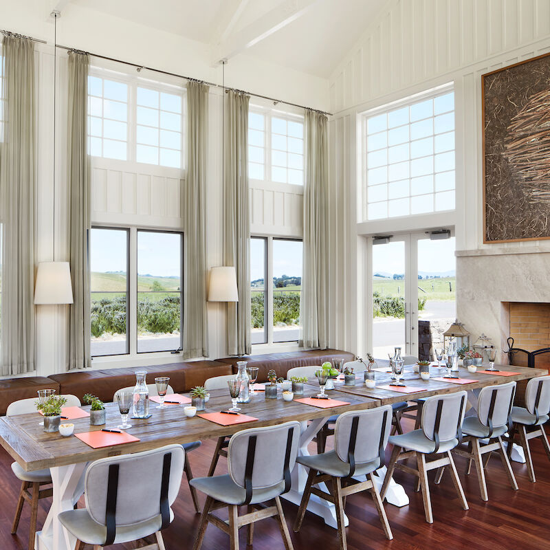 A well-lit dining room features a long table set for a meal, large windows with scenic views, high ceilings, and a lit fireplace with modern decor.