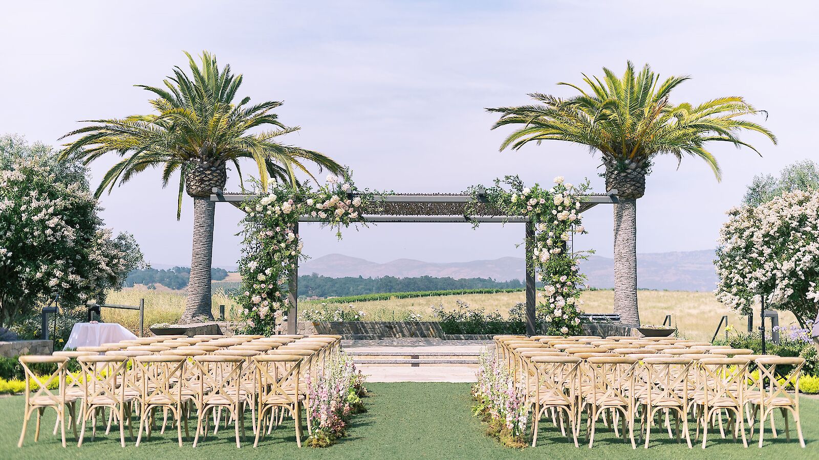 An outdoor wedding setup with chairs arranged facing a floral archway between two palm trees, against a scenic backdrop of fields and mountains.