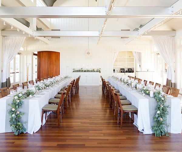 The image shows a bright, elegant dining room set for an event with two long tables adorned with white tablecloths and greenery, surrounded by wooden chairs.