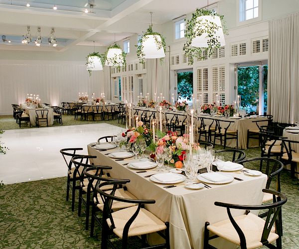 The image shows an elegant banquet hall set up for a formal event, with decorated tables, floral arrangements, and hanging lights.