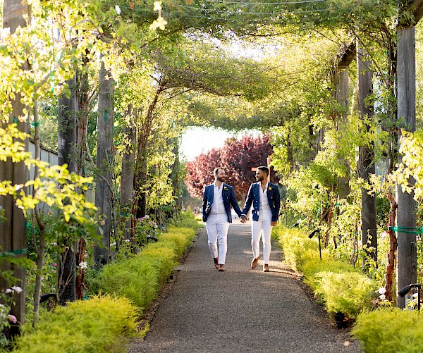 Two people are walking hand in hand down a garden pathway covered with green vines and foliage, creating a peaceful and romantic atmosphere.