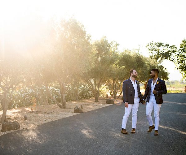 Two men are walking on a paved path under the sunlight. They both wear blazers and white pants, appearing to be engaged in conversation.