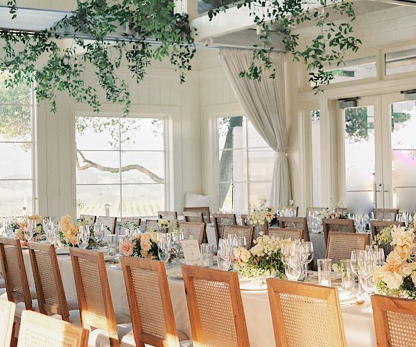The image shows an elegant indoor dining setup with long tables, wooden chairs, floral centerpieces, and lush green foliage hanging from the ceiling.