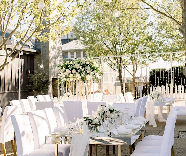 An outdoor dining setup with white chairs, a long table adorned with flowers, and draped fabric under a canopy of trees with hanging lights.