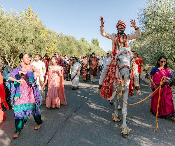 A joyful procession with a person riding a horse, surrounded by people in colorful attire, some playing instruments, on a sunny day.