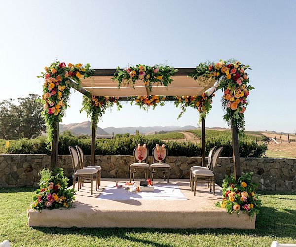 An outdoor wedding venue features a beautifully decorated altar with vibrant flowers, four chairs, and a table under a pergola.