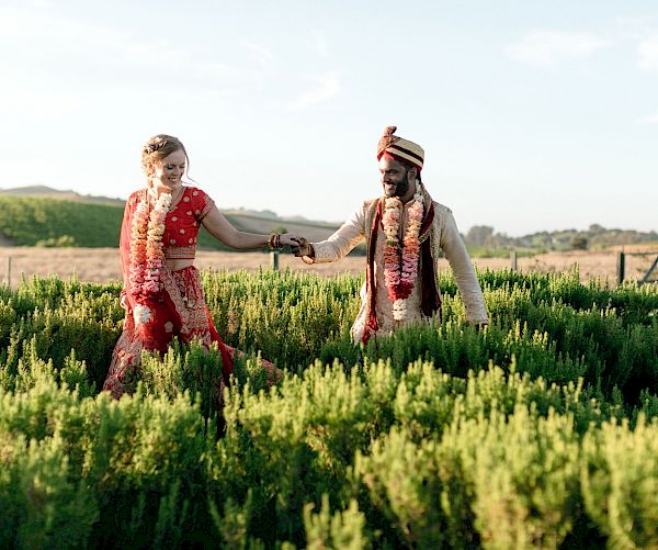 A couple in traditional attire walks through a green field, smiling and holding hands under a clear sky.