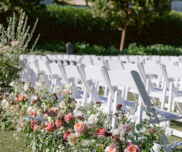 Rows of white folding chairs are arranged outdoors, adorned with floral arrangements at the aisle ends, under a clear sky with trees in the background.