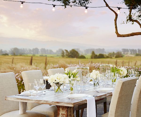 An elegantly set outdoor dining table with white flowers and candles overlooks a scenic, rural landscape under a string of lights at sunset.