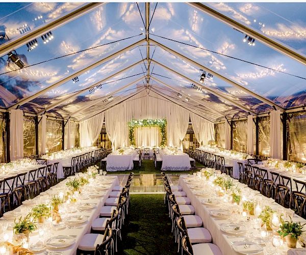 The image shows an elegantly decorated tent with long dining tables, adorned with white tablecloths, candles, and floral arrangements, under a clear roof.