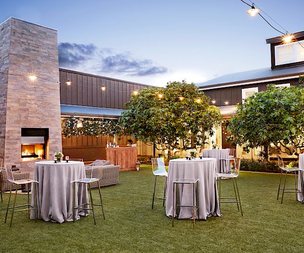 The image shows a well-decorated outdoor event space with round tables, chairs, a fireplace, trees, and string lights at dusk.