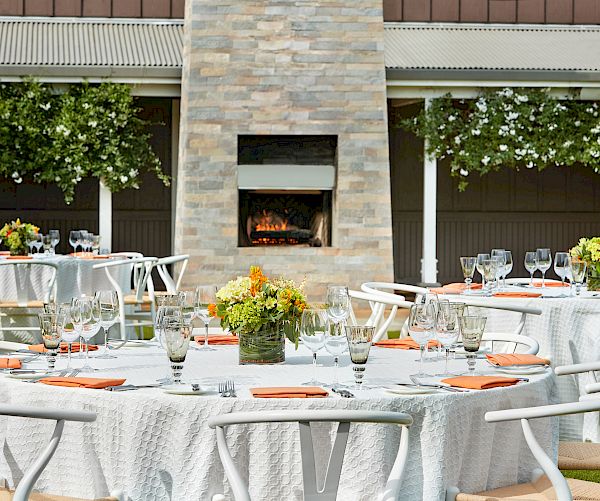 Outdoor event setup with round tables adorned with white tablecloths, flower centerpieces, and cutlery. A fireplace and hanging plants are visible.