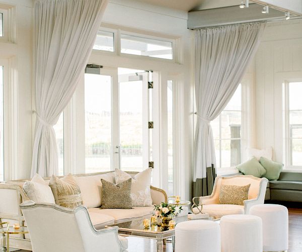 A stylish living room features white furniture, gold-accented stools, elegant drapes, and hardwood floors, with ample natural light from large windows.