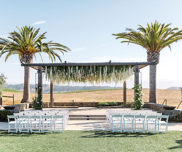 An outdoor wedding setup features white chairs facing an altar with hanging greenery, flanked by two palm trees under a clear sky.