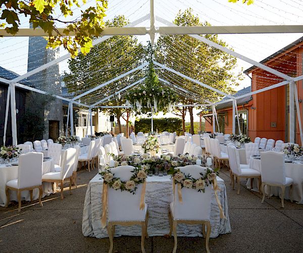 An elegant outdoor wedding reception setup under a transparent tent, with white chairs, tables adorned with floral arrangements, and overhead string lights.