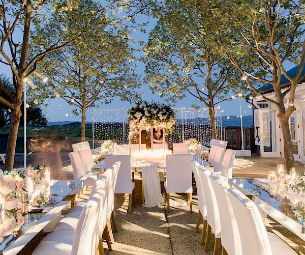 The image shows an elegant outdoor wedding setup with white chairs, floral decorations on tables, and fairy lights hanging from trees, creating a magical ambiance.