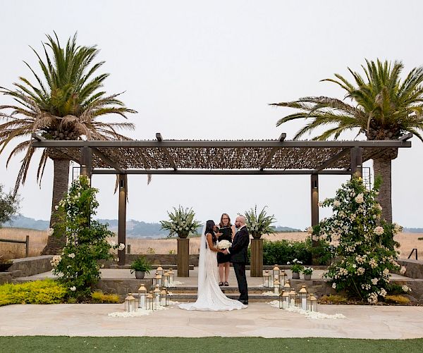 A couple is getting married outdoors beneath a pergola with two palm trees and flowers surrounding them, with candles lining the aisle.