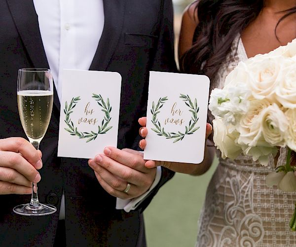 A wedding couple holding vow booklets and a bouquet, with the groom holding a glass of champagne in one hand and a booklet in the other hand.
