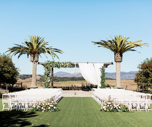 An outdoor wedding setup with white chairs arranged in rows on both sides of an aisle, a decorated arch, palm trees, and a scenic background.