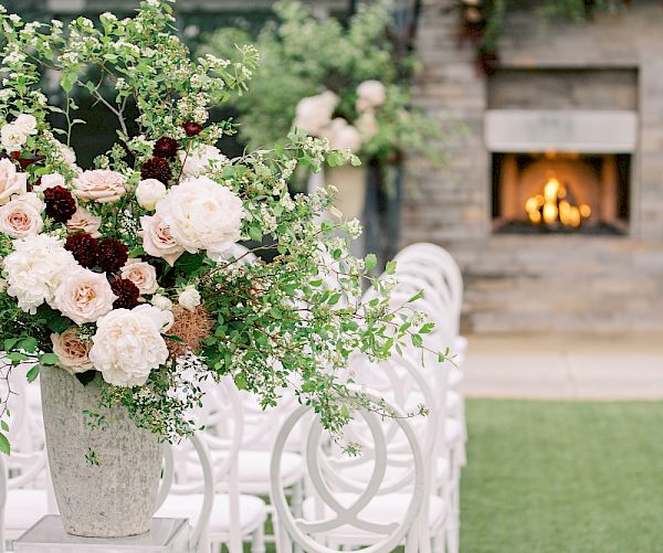 The image shows a tranquil outdoor setting with elegant white chairs, a large floral arrangement, and a lit fireplace in the background.