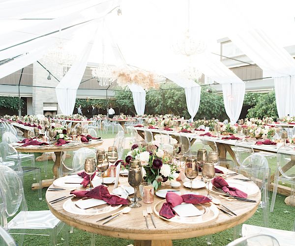The image shows a beautifully decorated outdoor event setup with round tables, elegant tableware, floral arrangements, and draped white fabric.