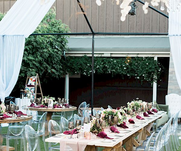 The image depicts an elegant outdoor event setup with long tables adorned with floral arrangements, chairs, and draped fabric, under a canopy.