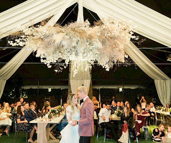 A couple is dancing under a decorative canopy at what looks like a wedding reception, with guests seated and watching in the background.
