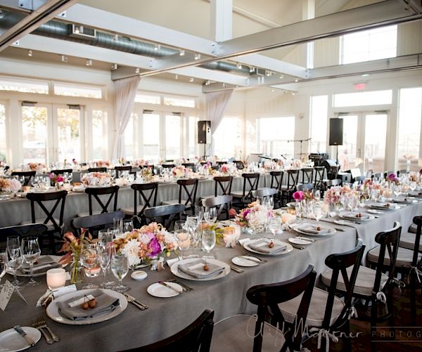 A beautifully set banquet hall with long tables adorned with colorful floral centerpieces, glassware, and dishes, ready for an elegant event.