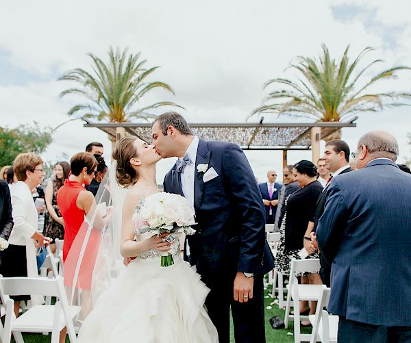 A couple is kissing at their wedding ceremony while surrounded by guests, with palm trees in the background.