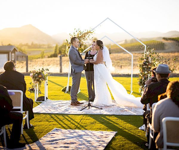 A couple is getting married outdoors with a small audience seated; the ceremony takes place with a scenic mountain backdrop and sunset lighting.