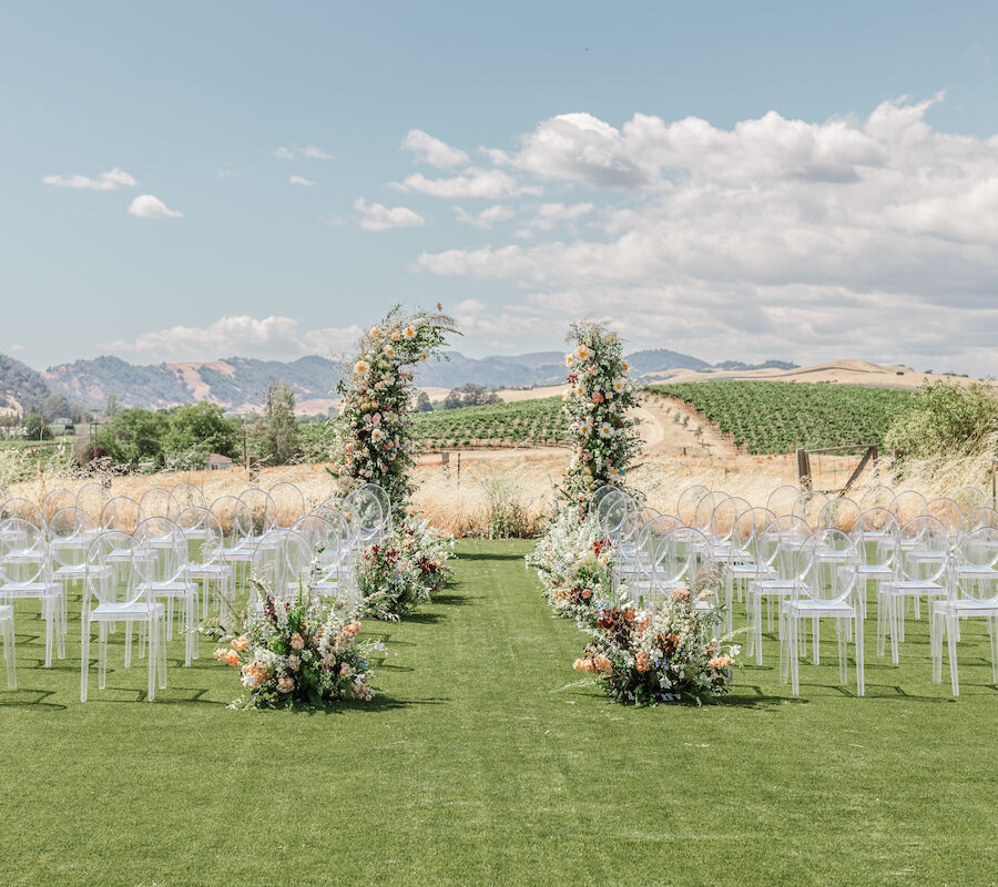 An outdoor wedding setup with clear chairs and a floral arch on green grass, surrounded by scenic hills and vineyards under a partly cloudy sky.
