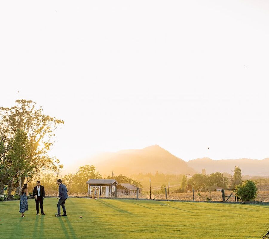 Three people stand on a lush, green field with mountains and a bright, setting sun in the background, enjoying a peaceful, picturesque scene.