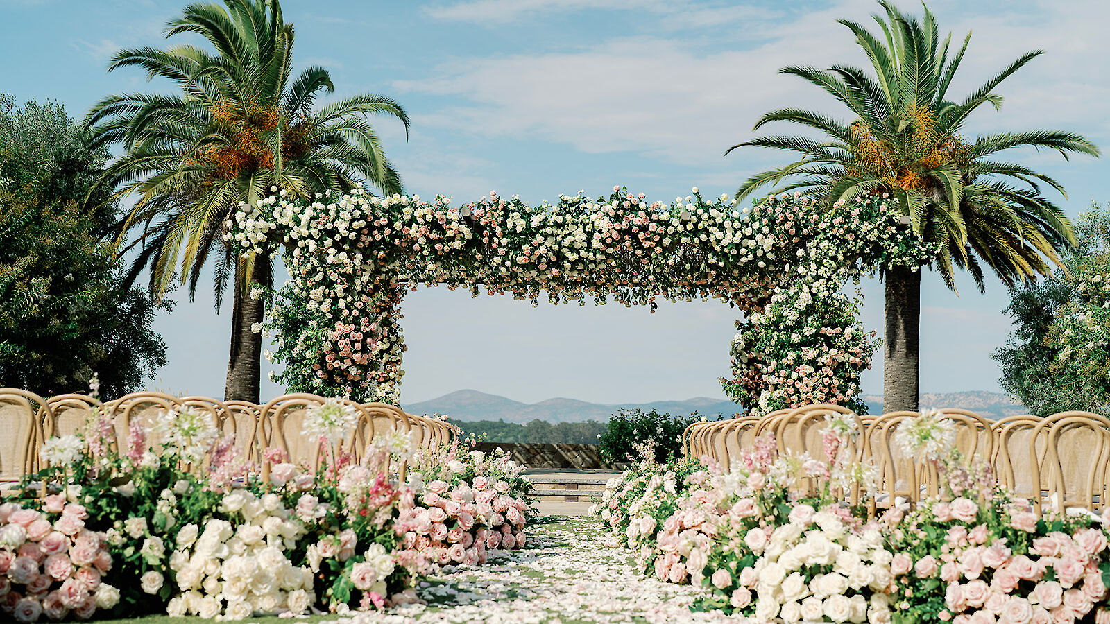 An outdoor wedding setup with floral arch, palm trees, and rows of chairs on a grassy aisle strewn with petals.